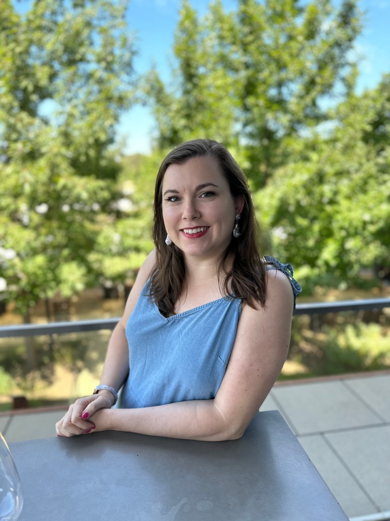 Photo of Lindsey Timpone, Lindsey is sitting at a table, with shoulder towards camera, smiling. Lindsey has brown hair to her shoulders, with a powder blue sleeveless top, with earrings and a bracelet.