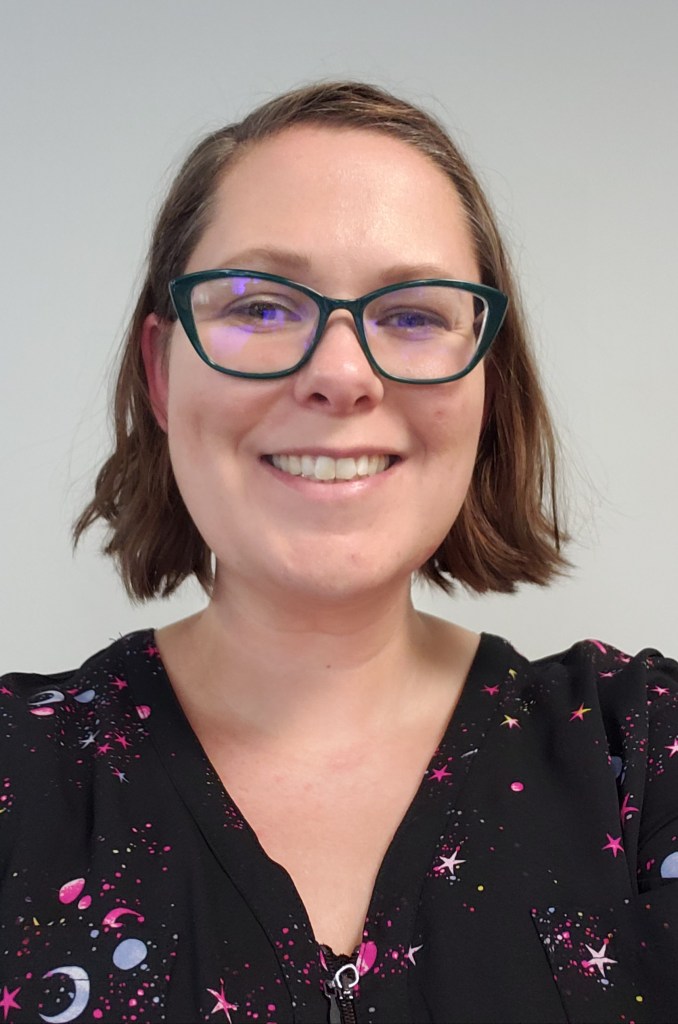A smiling light skinned woman with chin length brown hair, glasses, and a black v-neck blouse.