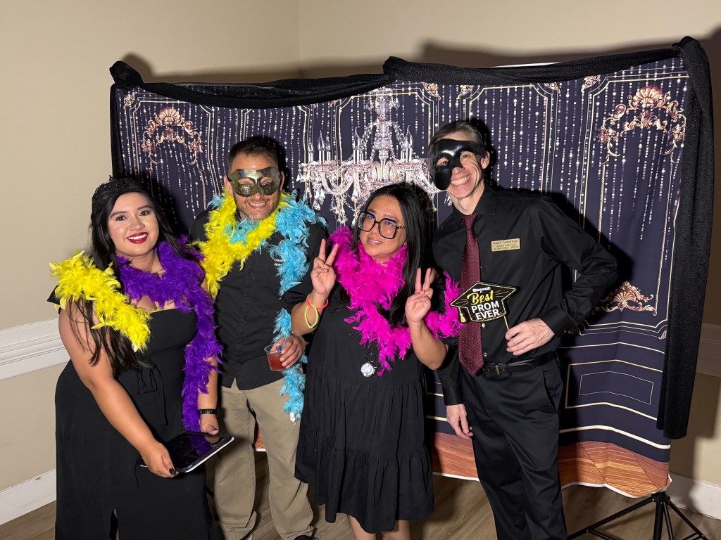 Carissa posing with three others during the free prom event in front of a photo background that has a classic blue wall with chandelier. All four are wearing mostly black with colorful feather boas. Two are wearing masks.