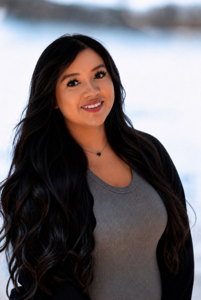 A woman, slightly turned in a professional pose, with medium brown skin and long dark wavy hair, wearing a dark open over coat with gray scoop neck shirt, and a simple necklace. She has a pleasant smile.