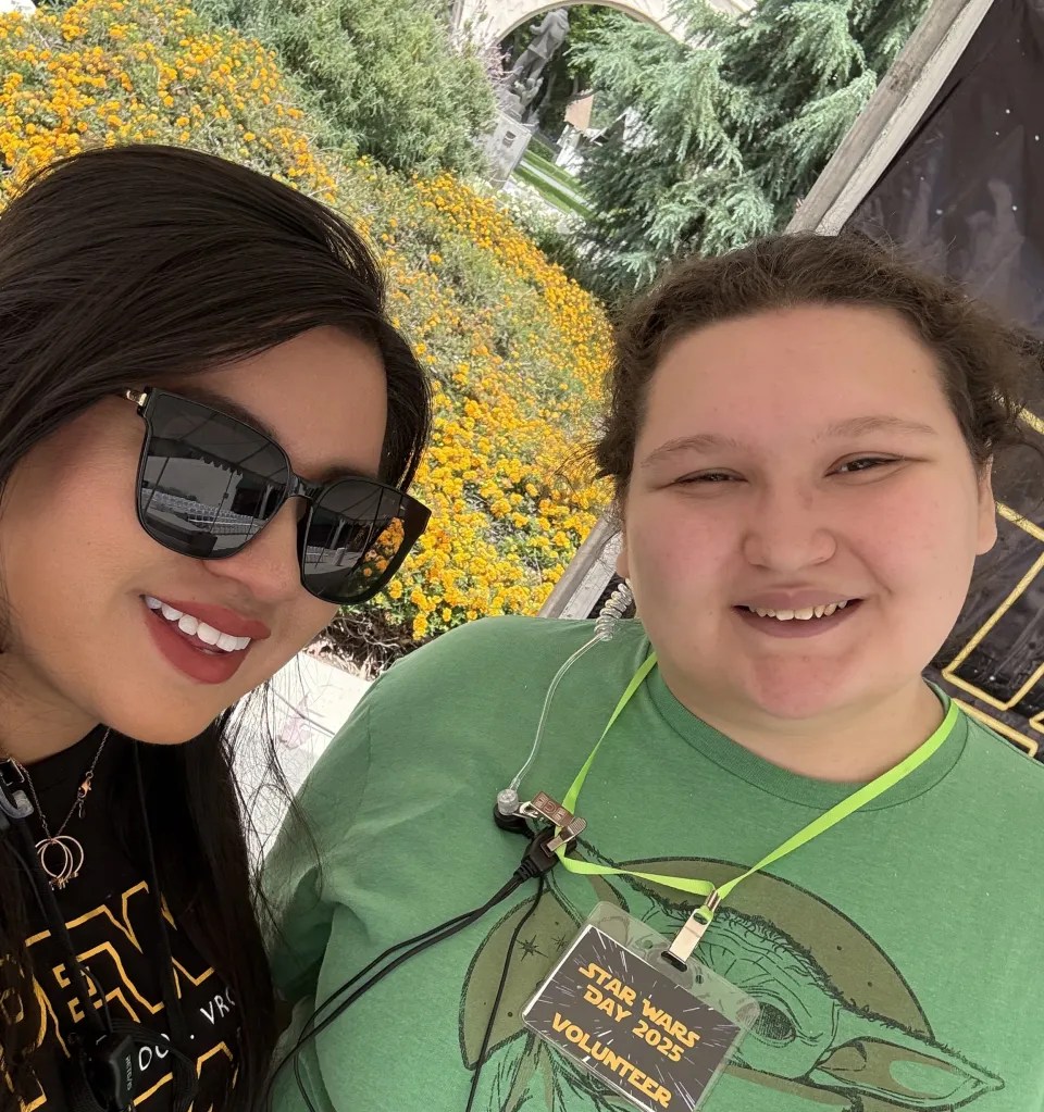 Carissa posing with a teen volunteer. Carissa is wearing black sunglasses and black shirt. The volunteer is has a light skin tone, brown hair that is pulled back and a green Yoda t-shirt. The volunteer is wearing a badge that says "Star Wars Day 2025 Volunteer." Both are smiling towards the camera.