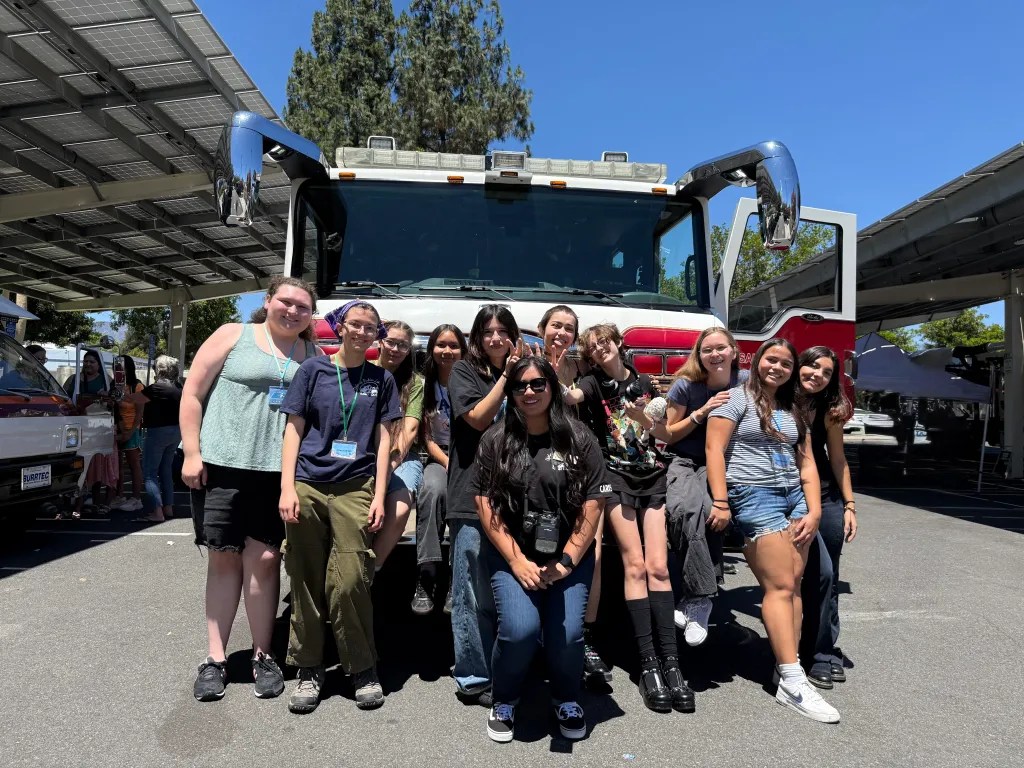 Carissa Barrera slightly crouched down in front of a group of nine teenagers posing around her, some putting up bunny ear fingers on Carissa, all smiling. The group is standing in front of an emergency vehicle in a parking lot during an event.